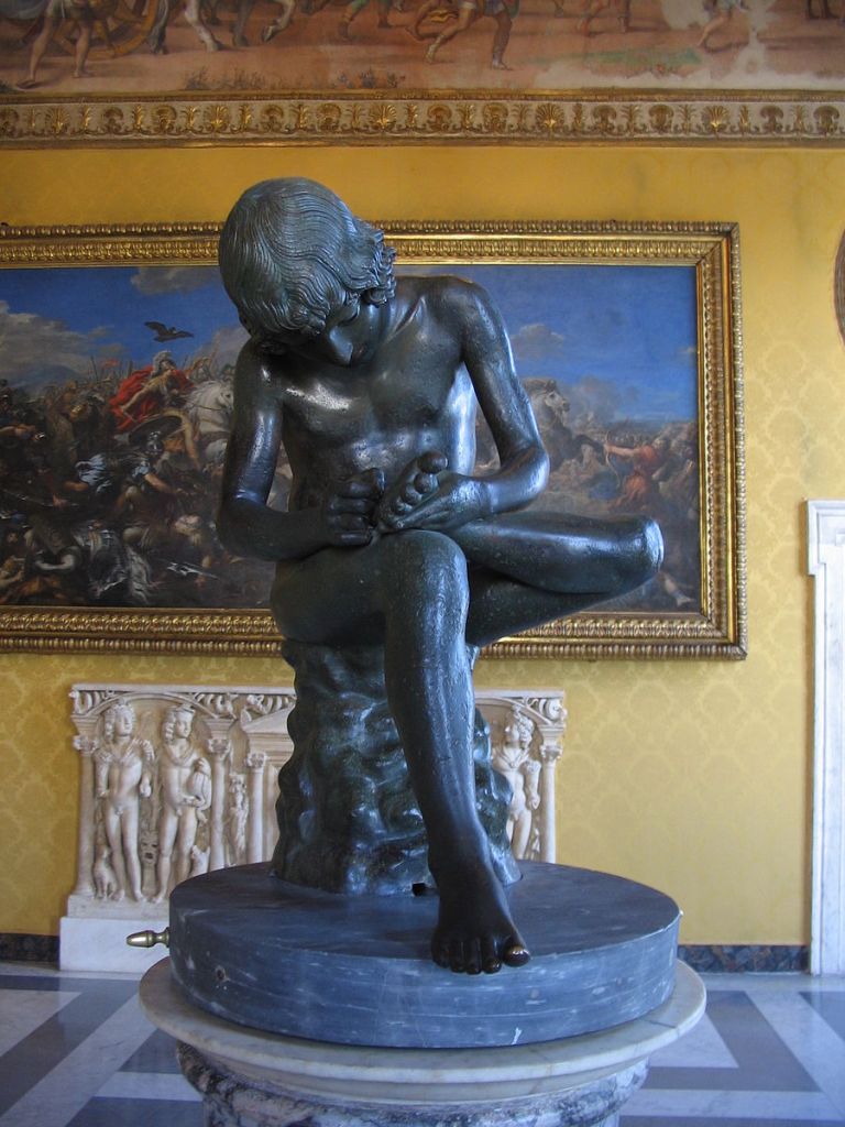 A photograph of a life-size bronze sculpture of a young teenage boy, seated, with chin-length who bends over his left foot, placed on his right knee, in order to pull a thorn from it with his right hand.